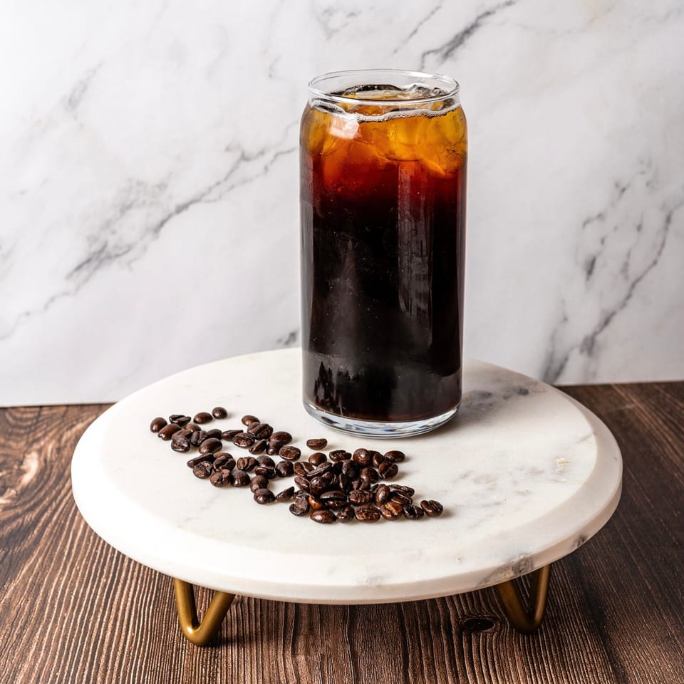 Iced coffee in a tall glass with coffee beans scattered on a marble stand.
