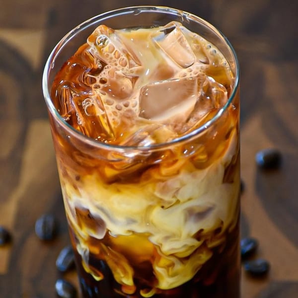 Iced coffee with cream swirling, ice cubes, and coffee beans scattered on a wooden surface.