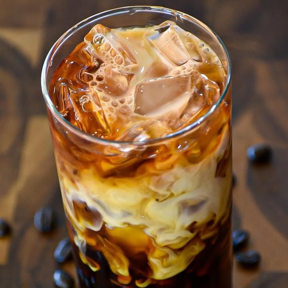 Iced coffee with cream swirling, ice cubes, and coffee beans scattered on a wooden surface.