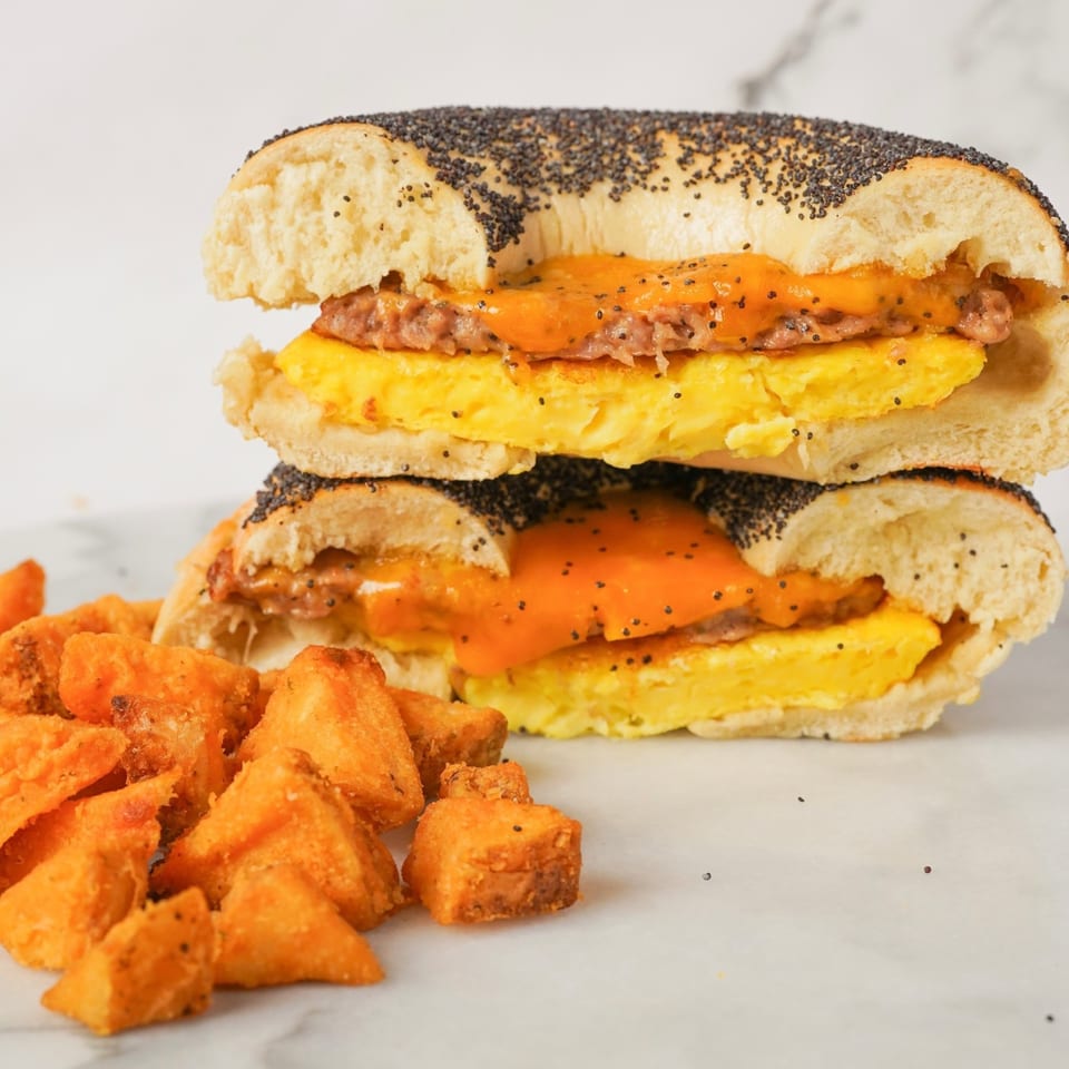 A poppy seed bagel sandwich with egg, sausage, and melted cheese, next to sweet potato fries.