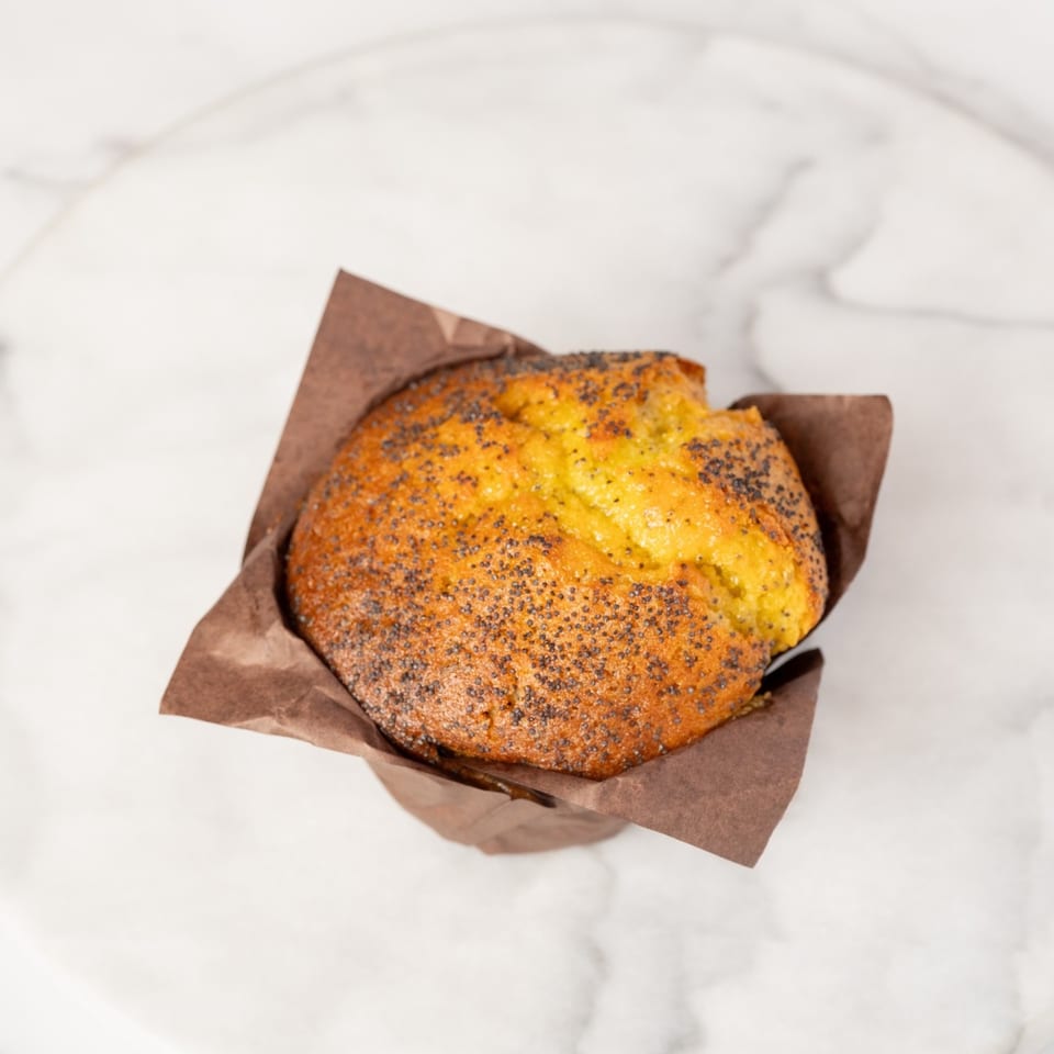 A golden-brown poppy seed muffin in a brown paper liner sits on a white marble surface.