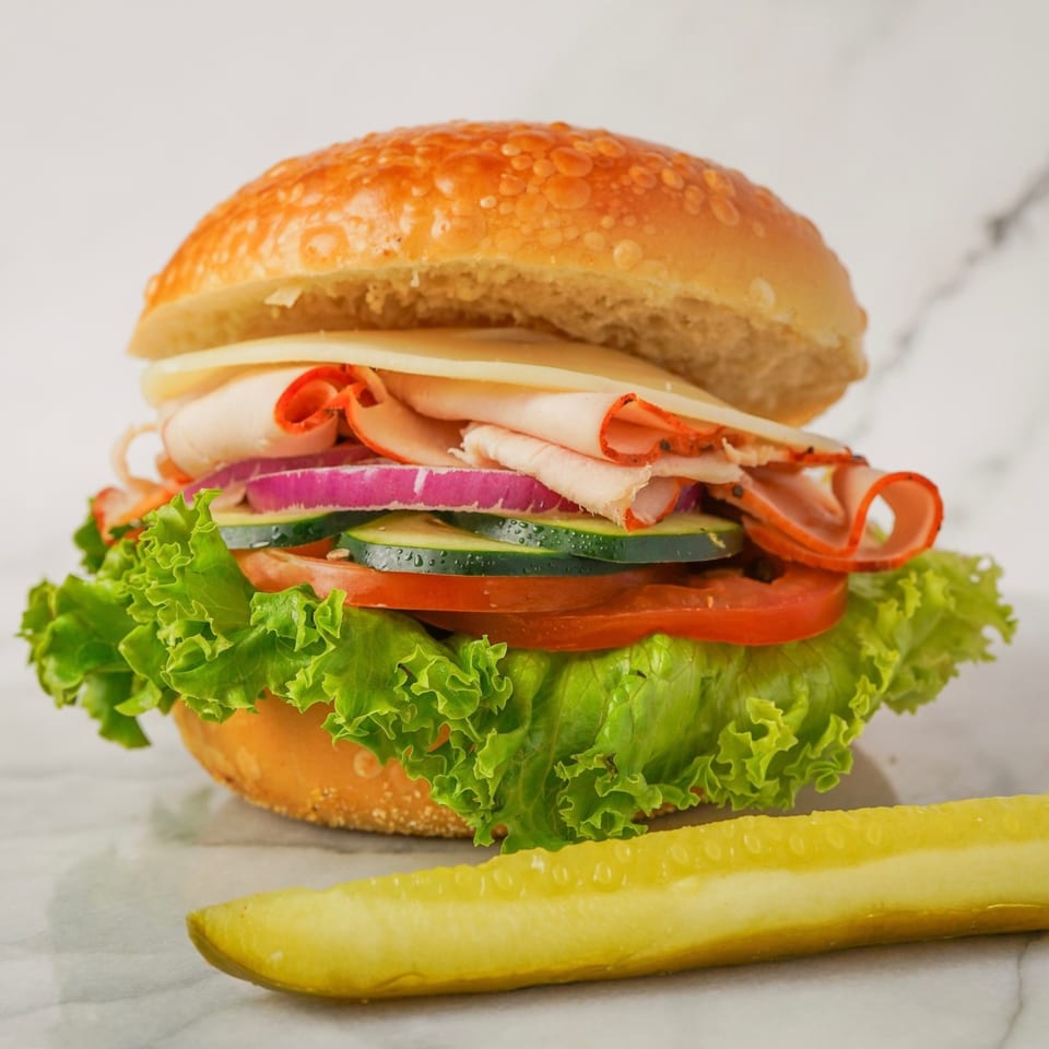 A Kokobelli Bagel Cafe bagel sandwich with turkey, cheese, lettuce, tomato, onion, and cucumber, with a pickle spear.